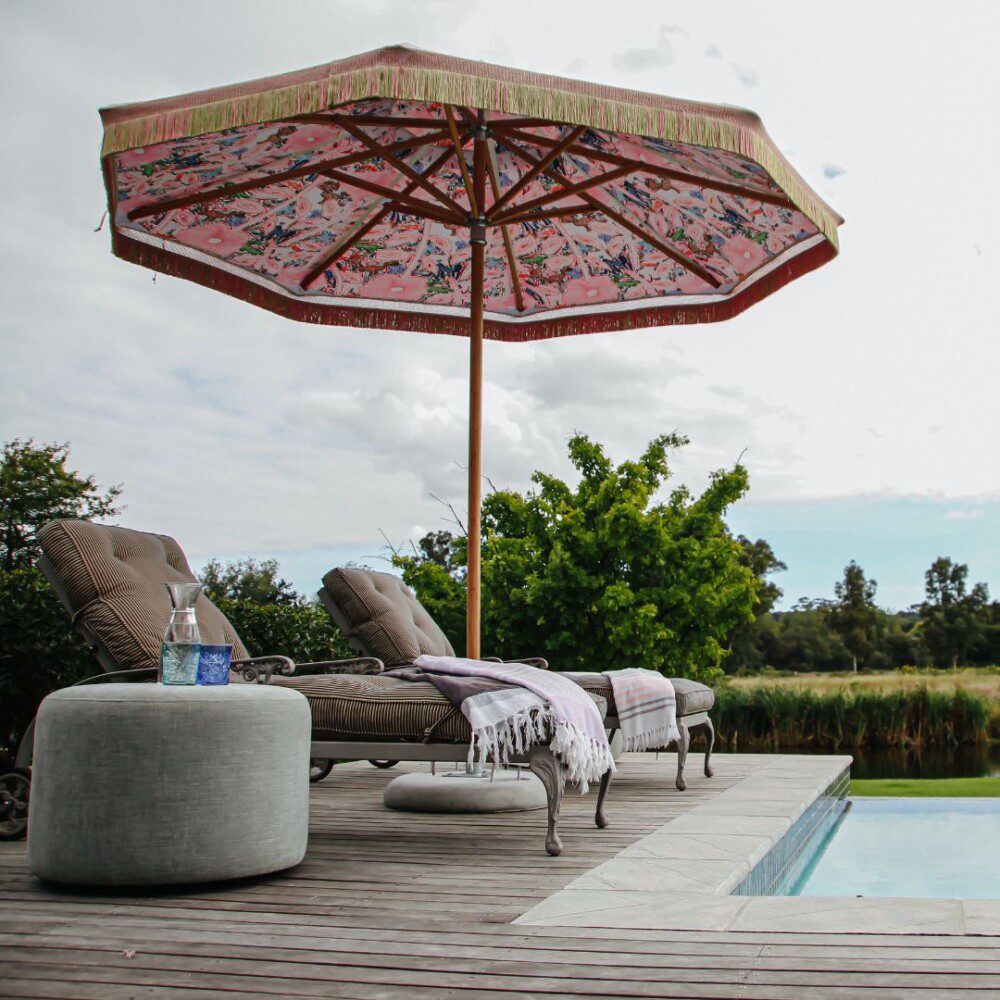 Special Montagu design parasol met houten frame, dubbele doekconstructie en kleurrijke binnenprint met franjes, geplaatst naast ligbedden op een luxe houten terras aan een zwembad in een groene tuinomgeving.